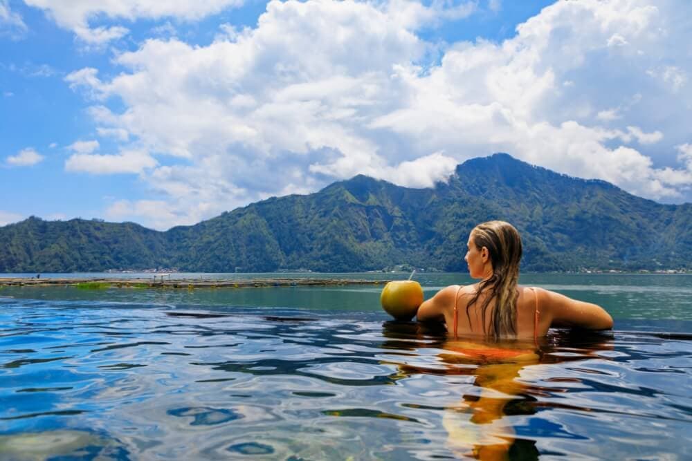 A person relaxes in water, holding a coconut, with mountains and a blue sky in the background.