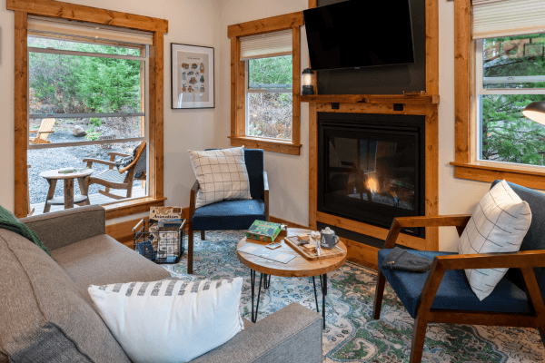 Cozy cabin sitting area with warm wood walls, a glowing fireplace, comfy chairs, and big windows looking out to the trees.