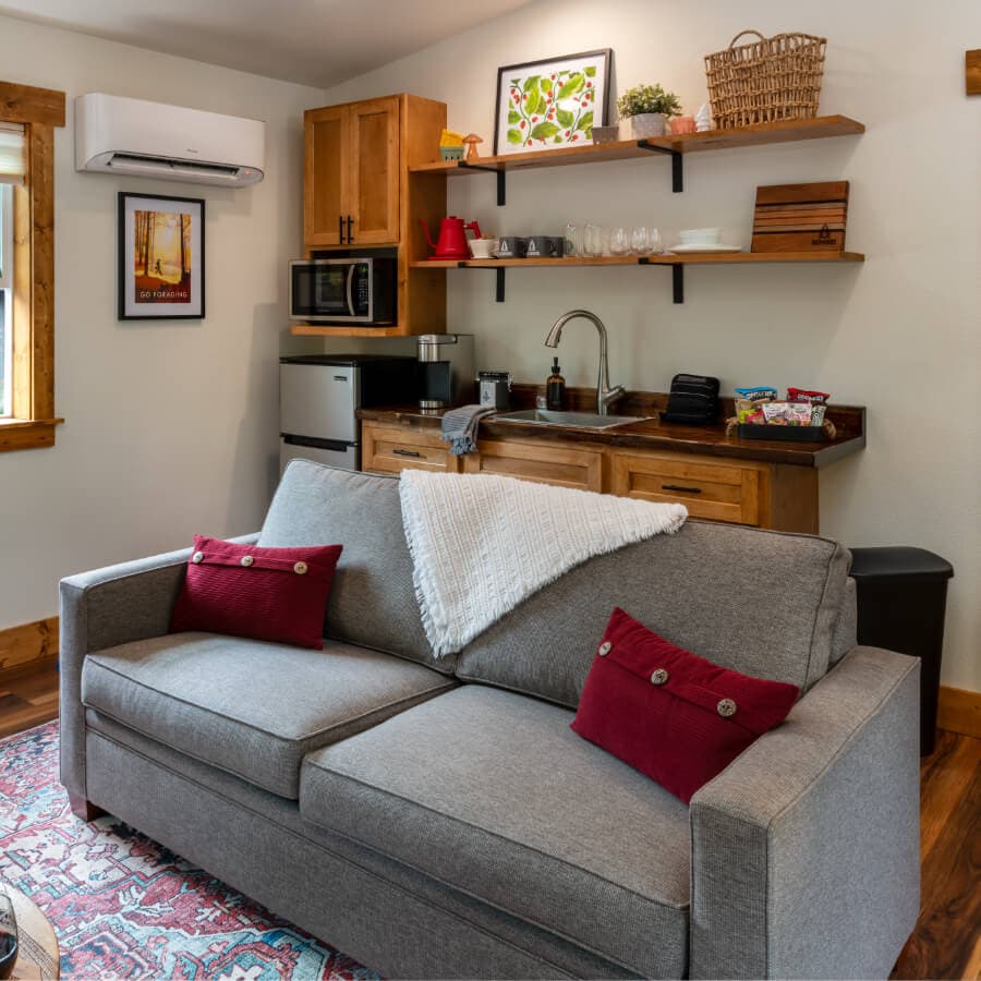 Cozy living space featuring a gray couch with red cushions, a wall-mounted air conditioner, and a kitchenette with wooden shelves.