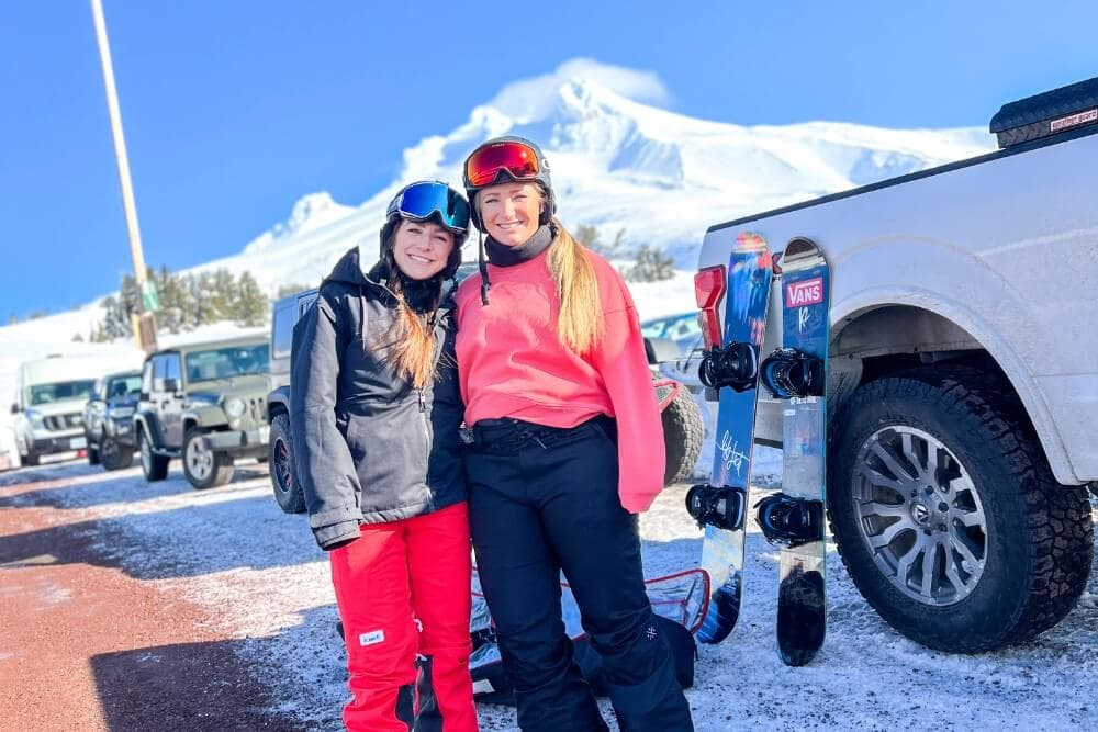 Two young women in ski gear pose together in a snowy parking area with a mountain backdrop.