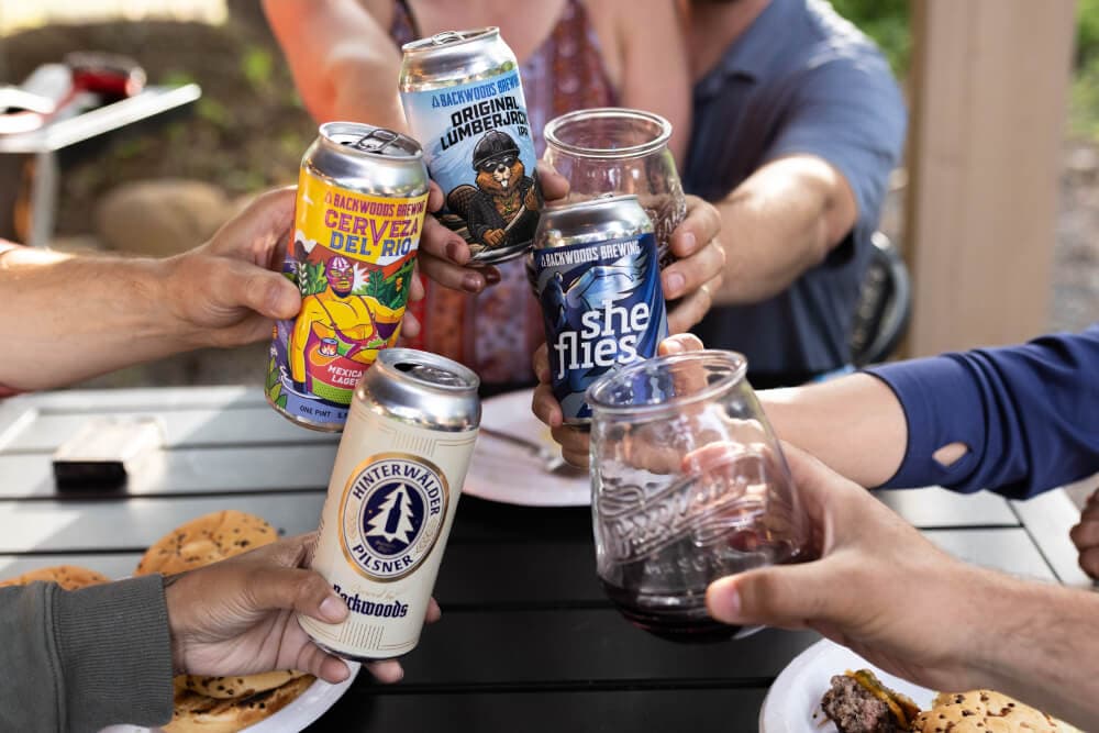 A group of hands raises various beer cans and glasses in a toast over a table with food.