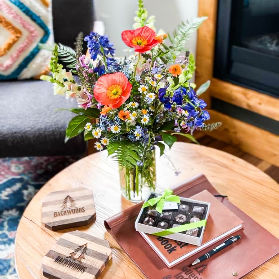 A vibrant bouquet of mixed flowers in a clear vase sits on a wooden table alongside coasters, a box of chocolates, and a notebook.