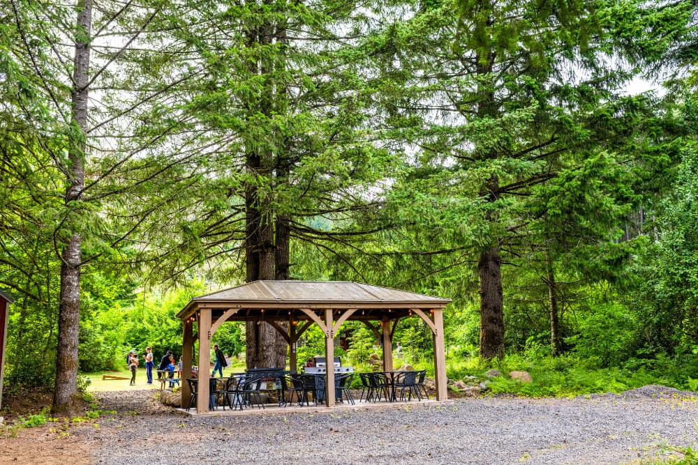 A wooden gazebo surrounded by tall trees and people walking in a lush green area.