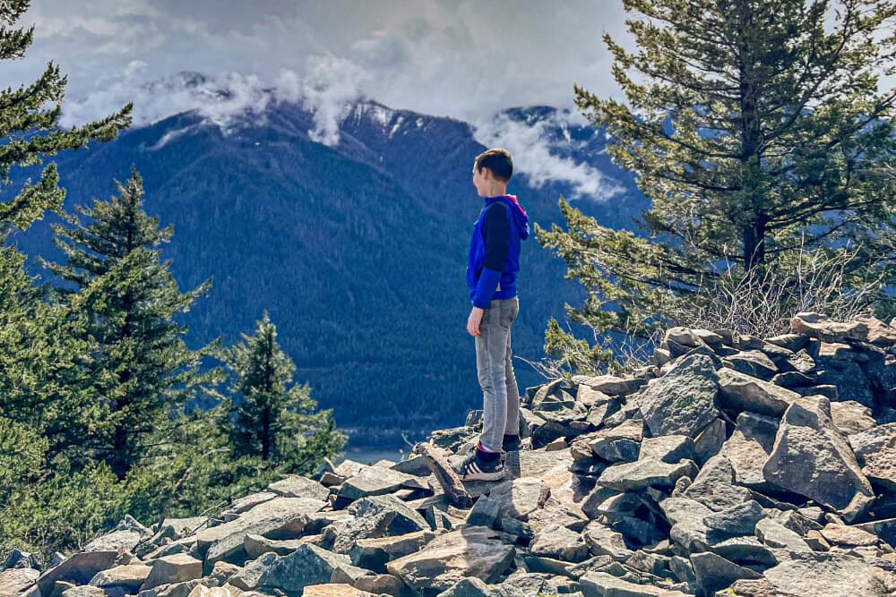 A child stands on a pile of rocks, overlooking a mountainous landscape with trees and clouds.
